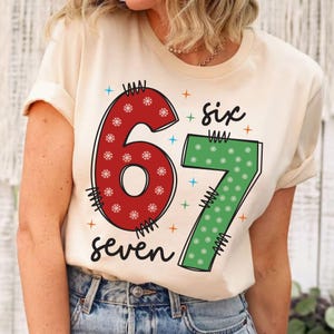 May include: A cream-colored t-shirt featuring the numbers "67" in red and green, decorated with snowflakes. The word "six" is above the number 7, and the word "seven" is below the numbers. The design includes blue and orange star accents.