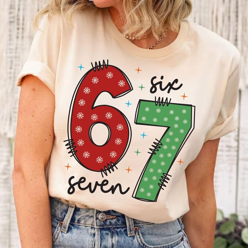 May include: A cream-colored t-shirt featuring the numbers "67" in red and green, decorated with snowflakes. The word "six" is above the number 7, and the word "seven" is below the numbers. The design includes blue and orange star accents.