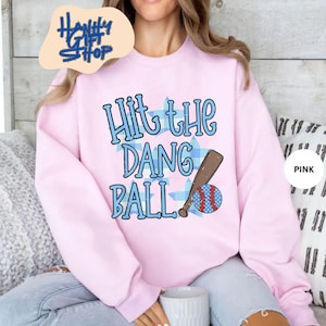 May include: Pink sweatshirt with the text "Hit the Dang Ball" in blue, along with a baseball bat and ball graphic. The baseball has red and blue stripes and a blue star pattern. The sweatshirt has a crew neck.