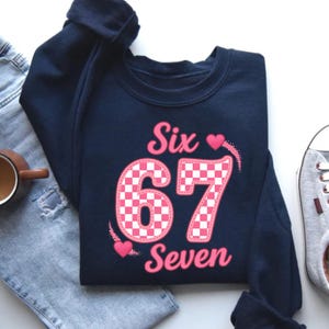 May include: Navy blue sweatshirt with the words "Six 67 Seven" in pink. The number "67" is in a pink and white checkered pattern. Two pink hearts are on the design.