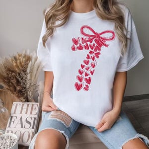 May include: White t-shirt featuring a red candy cane design made of hearts and a bow at the top. The candy cane design is drawn in a sketch style. The shirt is a short-sleeved crew neck.