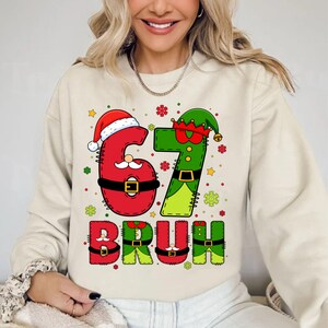 May include: Beige sweatshirt with a festive Christmas-themed graphic. The design features the numbers "67" dressed as Santa and an elf, with the word "BRUH" below. The letters and numbers are red and green with decorative accents.