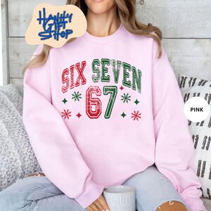 May include: A light pink sweatshirt with the text "SIX SEVEN 67" in red and green, with snowflake and diamond accents. The sweatshirt has a crew neck and long sleeves. The image also includes the text "Haniffy Shop".