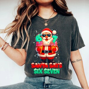 May include: Dark gray t-shirt featuring a cartoon Santa Claus wearing sunglasses and holding ice cream cones with the numbers 6 and 7. The text "SANTA SAYS SIX SEVEN" is printed below the Santa graphic.
