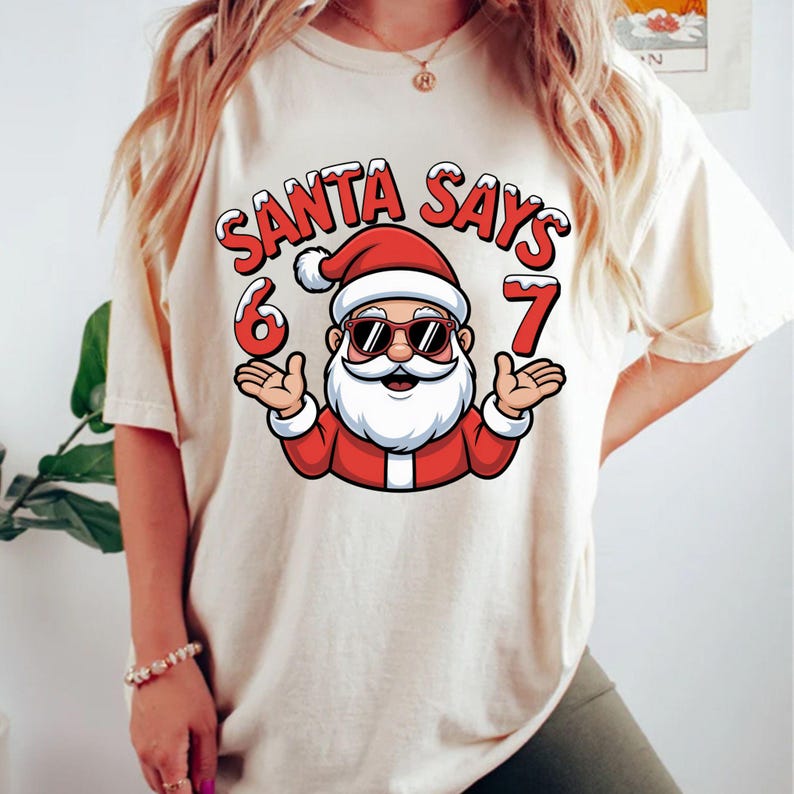 May include: Cream-colored t-shirt featuring a cartoon Santa Claus wearing sunglasses. The text "SANTA SAYS 6 7" is printed above the Santa graphic in red and white. The Santa illustration has a white beard and is wearing a red suit and hat.