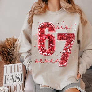 May include: Beige sweatshirt with the number "67" in red, filled with small red hearts. The word "six" is above the "6" and "seven" is below the "7", both in red script. A casual, comfortable style.