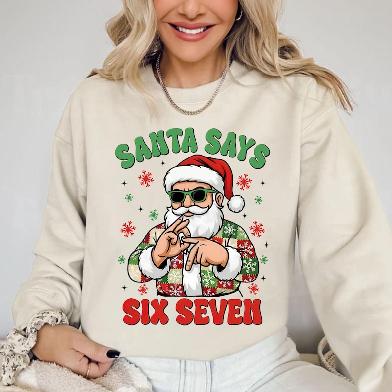 May include: Beige sweatshirt with a graphic of Santa Claus wearing sunglasses and a red hat. The Santa graphic has a white beard and is wearing a green, red, and white plaid jacket. The text "SANTA SAYS SIX SEVEN" is printed above and below the Santa graphic.