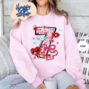 May include: Pink sweatshirt featuring a large number seven design with Valentine's Day themed graphics. The design includes hearts, a bow, a heart character wearing sunglasses, and the text "Hanify Gift Shop".