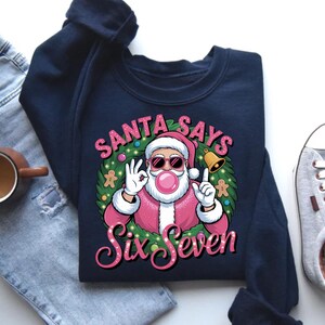 May include: Navy blue sweatshirt with a graphic of Santa Claus wearing sunglasses and blowing a bubblegum bubble. The text "SANTA SAYS Six Seven" is in a circular design around Santa.