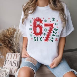 May include: White t-shirt with a red graphic featuring the numbers "67" and the words "SIX" and "SEVEN" in green. The design includes snowflakes and stars. The shirt is worn by a person in blue jeans.