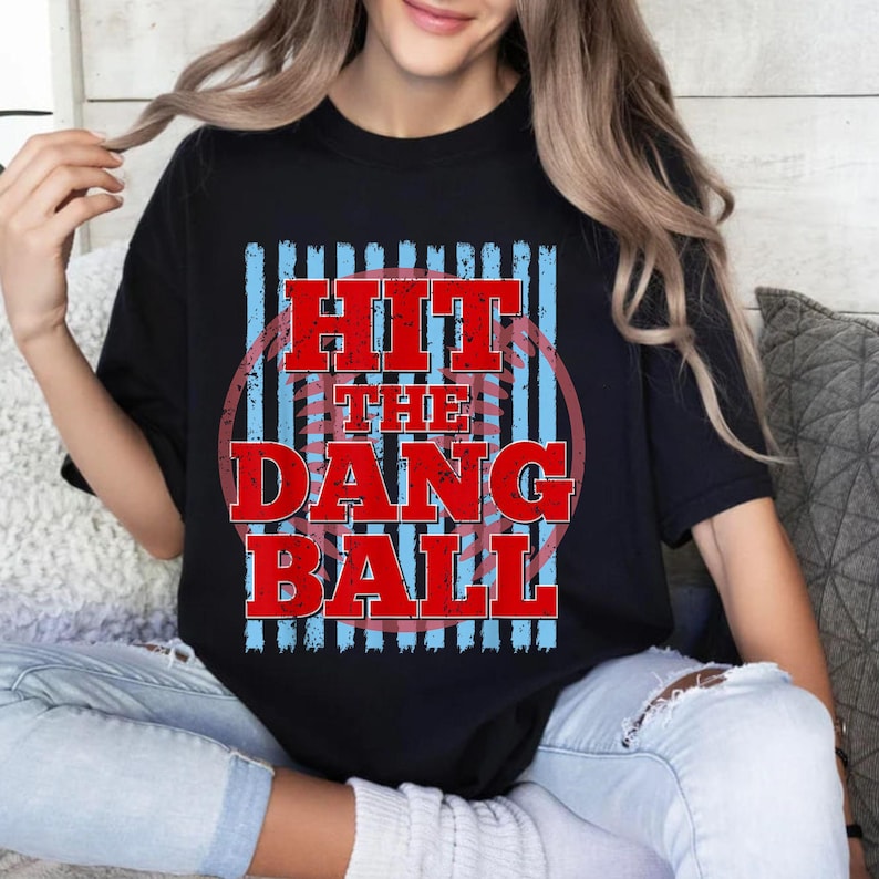 May include: Black t-shirt with the text "HIT THE DANG BALL" in red, distressed lettering. The design includes a baseball graphic and blue vertical stripes. The shirt is a casual style.