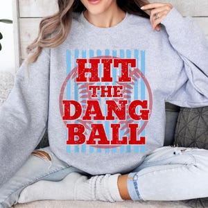 May include: A light gray sweatshirt with the text "HIT THE DANG BALL" in red, inside a red circle with blue stripes. The sweatshirt is a crew neck style.