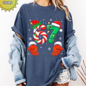 May include: Navy blue t-shirt with a festive Christmas design. The number "67" is featured, with the "6" designed as a candy cane and the "7" in green with a Santa hat. Snowflake and hand graphics complete the holiday theme.