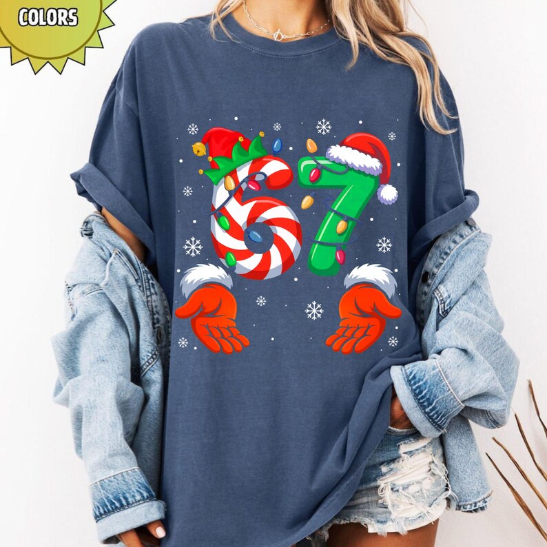 May include: Navy blue t-shirt with a festive Christmas design. The number "67" is featured, with the "6" designed as a candy cane and the "7" in green with a Santa hat. Snowflake and hand graphics complete the holiday theme.