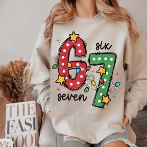 May include: Beige sweatshirt with a festive design featuring the numbers "6" and "7" in red and green, adorned with string lights and stars. The word "six" and "seven" are written in black.