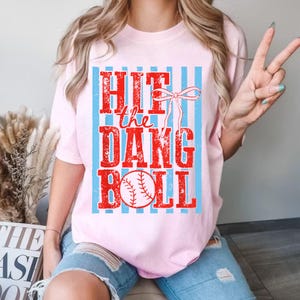 May include: A light pink t-shirt with the text "Hit the Dang Ball" in red, with a baseball graphic. The text is set against a background of light blue vertical stripes. The shirt has short sleeves.