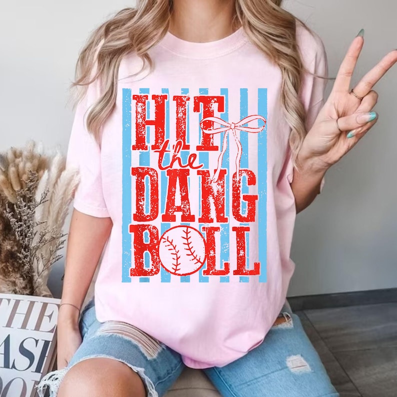 May include: A light pink t-shirt with the text "Hit the Dang Ball" in red, with a baseball graphic. The text is set against a background of light blue vertical stripes. The shirt has short sleeves.