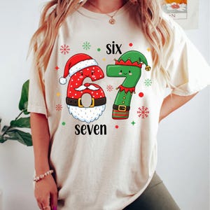 May include: Cream-colored t-shirt featuring a festive design with the numbers "6" and "7" dressed as Santa and an elf. The number "6" is red with white polka dots and a Santa hat. The number "7" is green with an elf hat. The word "six" and "seven" are also printed on the shirt.