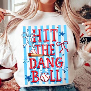 May include: White sweatshirt with a baseball-themed graphic. The design features the text "HIT THE DANG BALL" in red, with a baseball and bat, bows, and blue stars on a blue striped background.