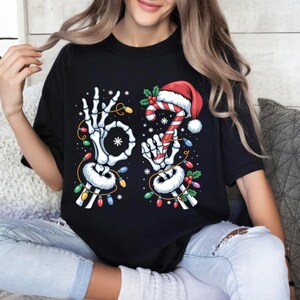 May include: Black t-shirt featuring a skeleton hand making an "okay" sign and another holding a candy cane with a Santa hat. The design includes Christmas lights and snowflakes, creating a festive holiday theme.