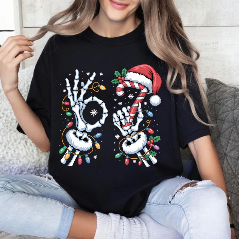 May include: Black t-shirt featuring a skeleton hand making an "okay" sign and another holding a candy cane with a Santa hat. The design includes Christmas lights and snowflakes, creating a festive holiday theme.