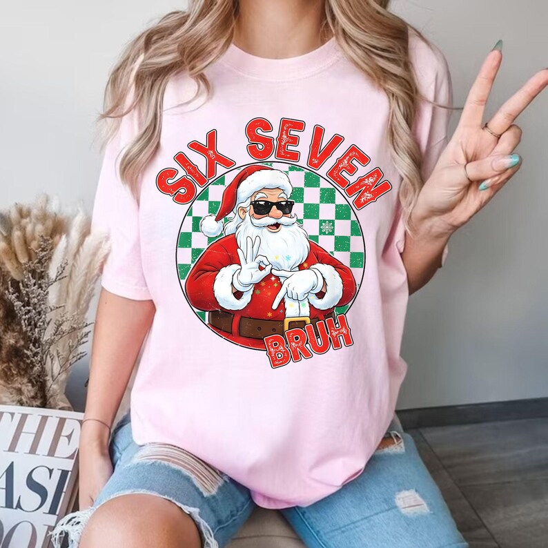 May include: A light pink t-shirt featuring a graphic of Santa Claus wearing sunglasses and giving a peace sign. The text "SIX SEVEN BRUH" is printed above and below the Santa graphic. The Santa graphic is inside a green and white checkered circle.