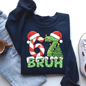 May include: Navy blue sweatshirt with a festive Christmas design. The graphic features the numbers "67" decorated with candy cane and Christmas lights, topped with Santa hats. The word "BRUH" is in green with white polka dots.