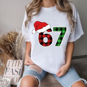 May include: White t-shirt featuring the number "67" in a festive design. The "6" is red and black plaid, topped with a Santa hat. The "7" is green and black plaid. A fun, holiday-themed graphic tee.