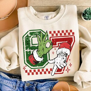 May include: Cream-colored sweatshirt with a festive graphic. The design features the number 87 in green and red, with a Grinch-like hand making an "OK" gesture and a Santa-like hand pointing down. Red and white checkered accents and star details complete the holiday theme.
