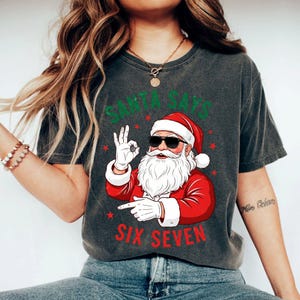 May include: Dark gray t-shirt featuring a graphic of Santa Claus wearing sunglasses and making an 'okay' hand gesture. The text "SANTA SAYS" is above the Santa graphic, and "SIX SEVEN" is below.