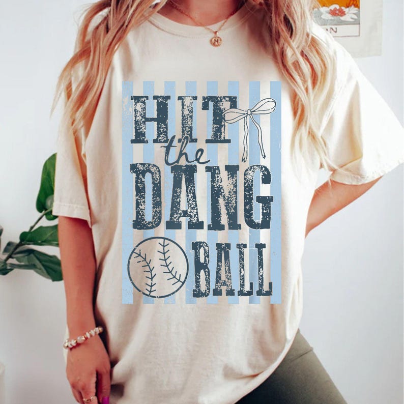 May include: Cream-colored t-shirt with the phrase "HIT the DANG BALL" in distressed navy blue lettering. A baseball graphic is below the text. Light blue vertical stripes and a white bow accent the design.