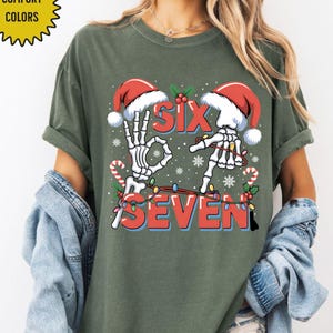 May include: Olive green t-shirt with a festive design. The graphic features skeleton hands forming the numbers "six" and "seven" with Santa hats, candy canes, and string lights. The text "COMFORT COLORS" is in the upper left corner.