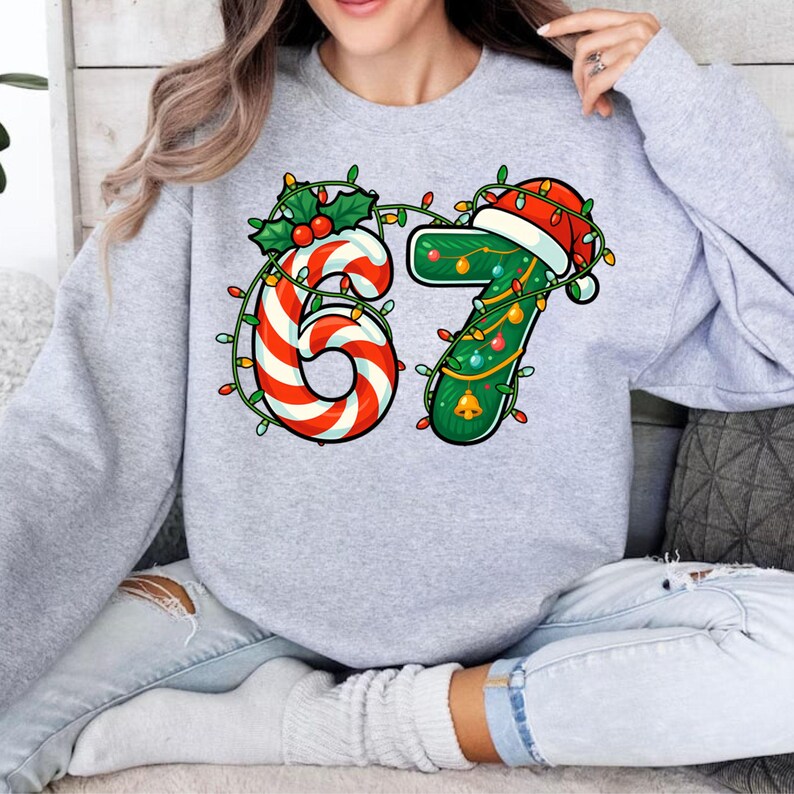 May include: A light gray sweatshirt featuring a festive design with the number "67". The "6" is a red and white candy cane design, while the "7" is green with a Santa hat and Christmas lights. A fun holiday-themed top.