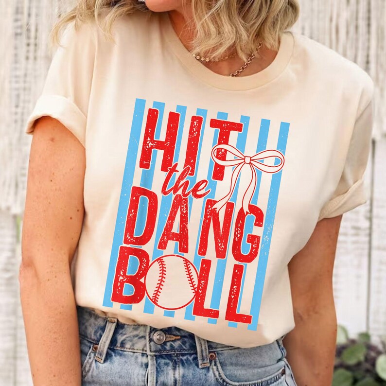 May include: Cream-colored t-shirt with the phrase "HIT the DANG BALL" in red, with a baseball graphic. The text is set against a backdrop of blue vertical stripes and a white bow.