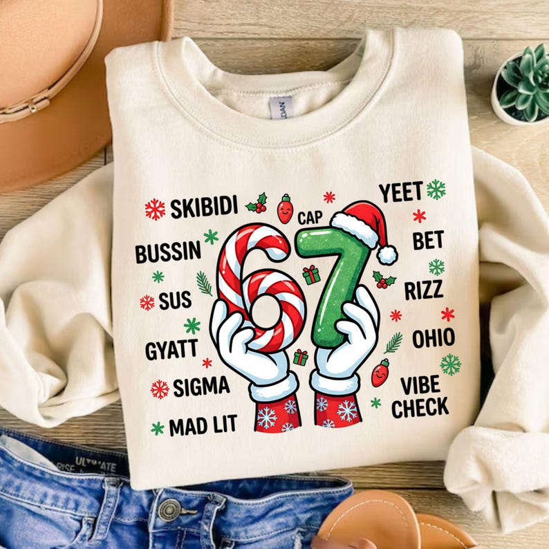 May include: Cream-colored sweatshirt with a festive design. The graphic features the number "67" in a candy cane and Santa hat design, surrounded by holiday-themed text and snowflakes. The sweatshirt is folded on a wooden surface.