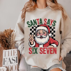 May include: Beige sweatshirt with a graphic of Santa Claus giving the peace sign. The text "SANTA SAYS" is above Santa, and "SIX SEVEN" is below. The background is a black and white checkered pattern.