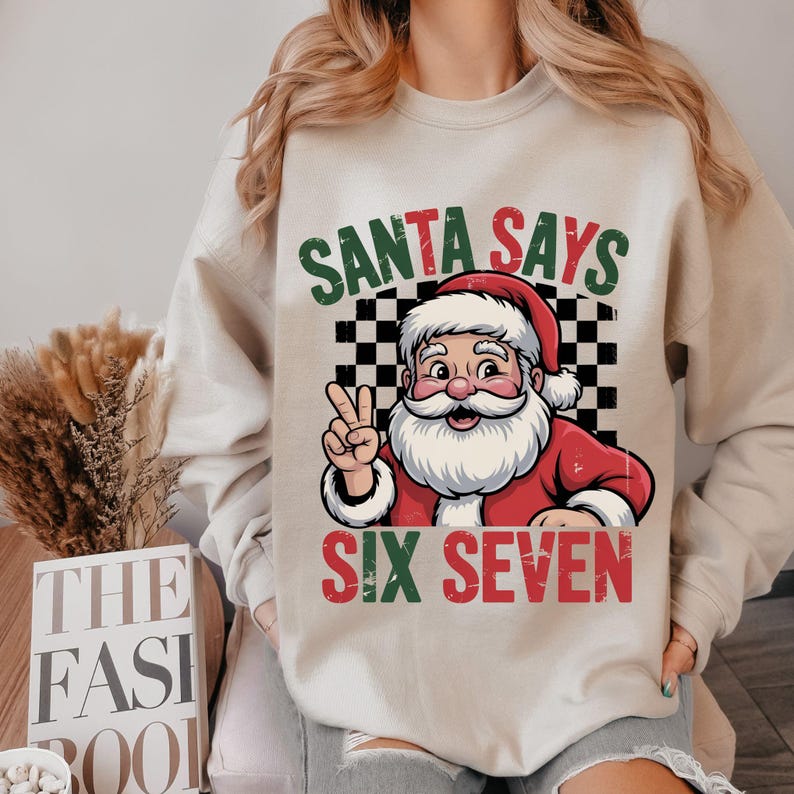 May include: Beige sweatshirt with a graphic of Santa Claus giving the peace sign. The text "SANTA SAYS" is above Santa, and "SIX SEVEN" is below. The background is a black and white checkered pattern.