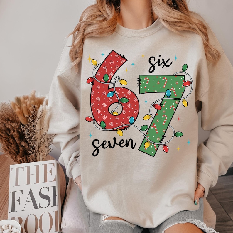 May include: Beige sweatshirt with a festive design featuring the numbers "6" and "7" in red and green, adorned with Christmas lights. The word "Six" is above the numbers, and "Seven" is below.