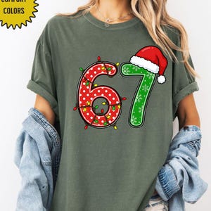 May include: A green t-shirt with the numbers "67" in a Christmas theme. The number "6" is red with white polka dots and wrapped in Christmas lights. The number "7" is green with snowflakes and a Santa hat.