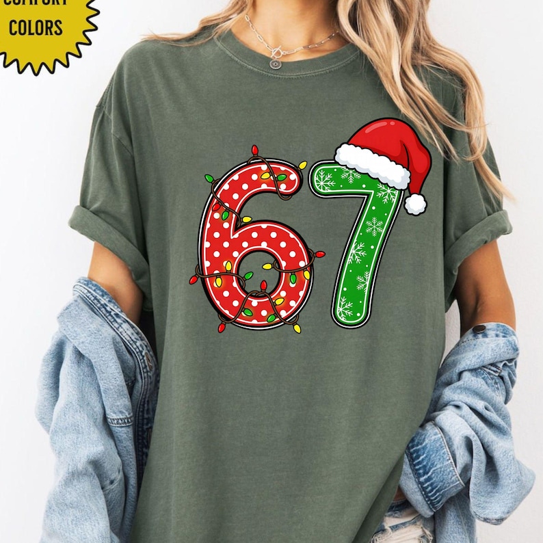 May include: A green t-shirt with the numbers "67" in a Christmas theme. The number "6" is red with white polka dots and wrapped in Christmas lights. The number "7" is green with snowflakes and a Santa hat.