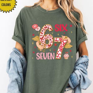 May include: Green t-shirt with the words "SIX SEVEN" in pink. The number 6 and 7 are decorated with a heart pattern. The shirt has a graphic of Cupid with wings and a cupcake.
