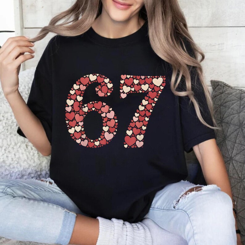 May include: Black t-shirt with the number "67" in a heart-shaped design. The hearts are in shades of red and cream. The shirt is worn by a person wearing blue jeans.