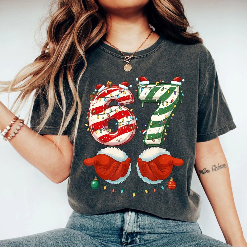 May include: Dark gray t-shirt featuring a festive Christmas design. The number "67" is decorated with candy cane stripes and green stripes, adorned with Santa hats and string lights. Below are red gloved hands holding ornaments.