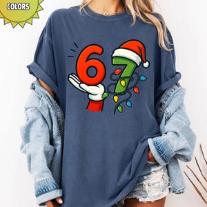May include: Navy blue t-shirt featuring the numbers 6 and 7 in a festive design. The number 6 is red and held by a gloved hand, while the number 7 is green and adorned with a Santa hat and Christmas lights. The word "COLORS" is in a yellow sunburst.