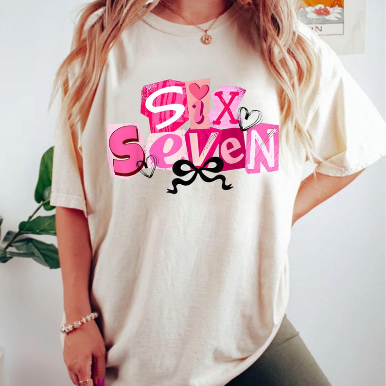 May include: Cream-colored t-shirt featuring the words "Six Seven" in a pink and white block letter design. The design includes pink hearts and a black bow. The shirt is a casual, oversized style.