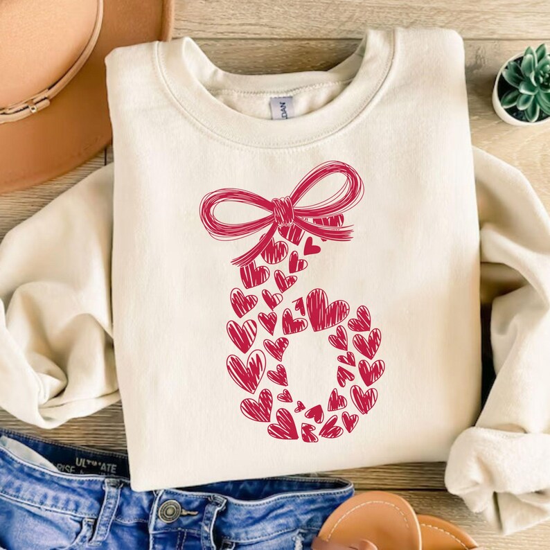May include: Cream-colored sweatshirt featuring a large red bow at the top and a number six formed from numerous red hearts. The sweatshirt is folded on a wooden surface, with a pair of blue jeans visible in the foreground.