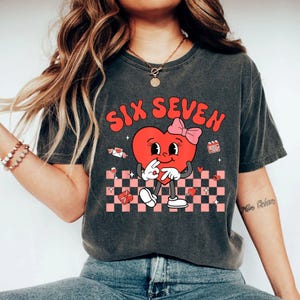 May include: Dark gray t-shirt featuring a retro-style graphic of a heart character with a pink bow, hands making a peace sign, and the words "SIX SEVEN" in red. The design includes a pink and white checkered pattern.