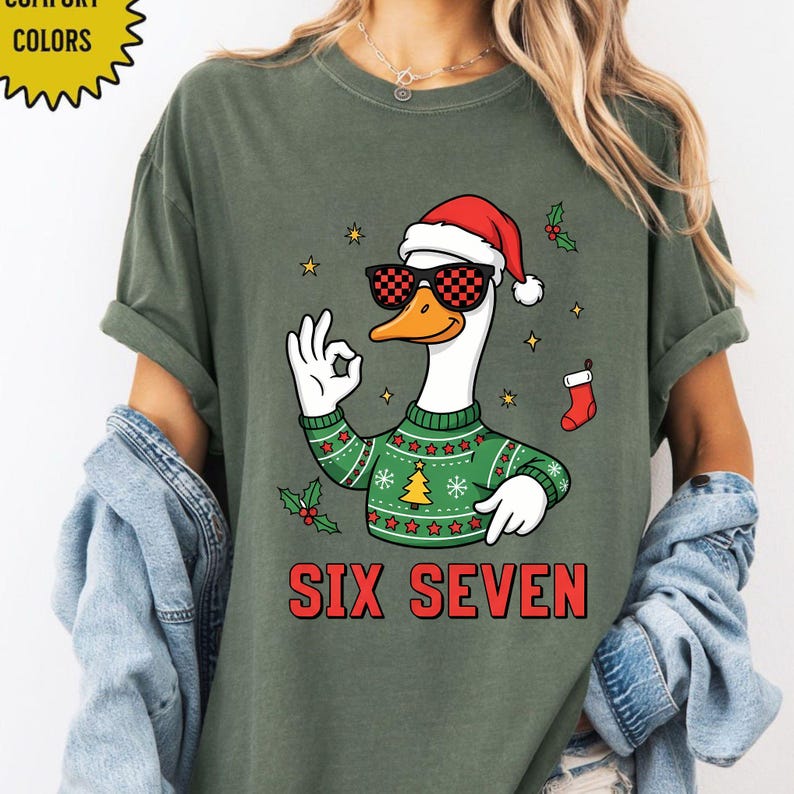 May include: Green t-shirt featuring a cartoon goose wearing sunglasses and a Santa hat, giving an "okay" hand gesture. The goose is wearing a green sweater with a Christmas tree design. The text "SIX SEVEN" is printed in red.