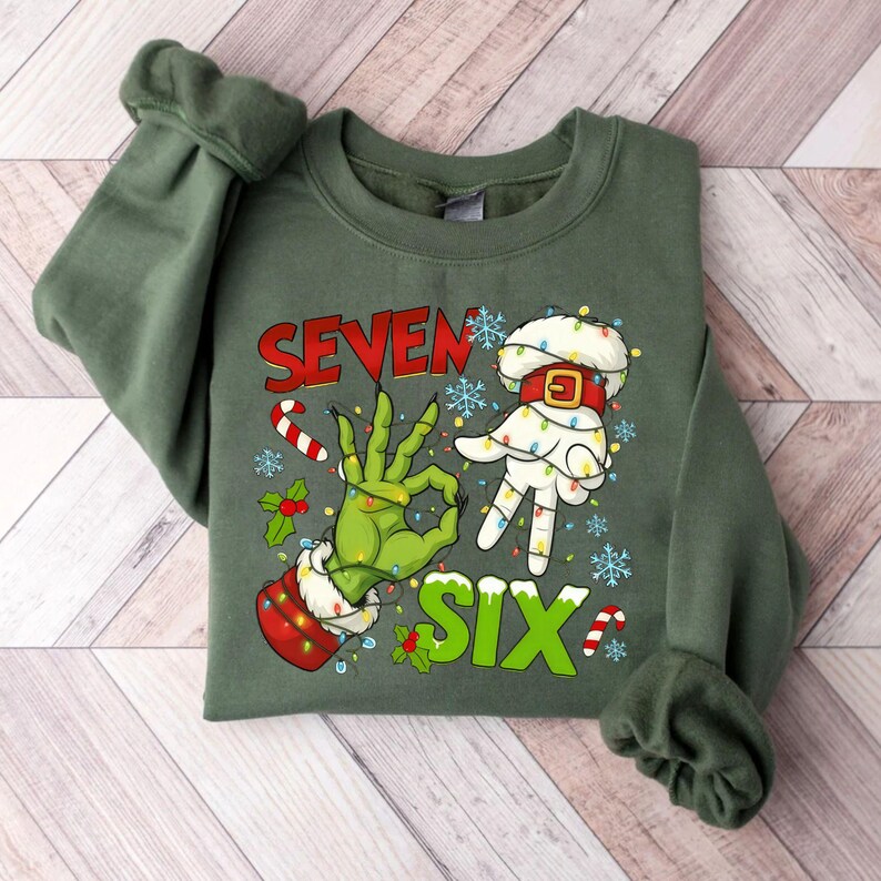 May include: Green sweatshirt with a festive design. The graphic features the words "SEVEN SIX" in red and green, along with illustrations of hands making hand gestures. The hands are decorated with Christmas lights and candy canes.