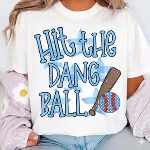 May include: White t-shirt with the text "Hit the Dang Ball" in blue, with a baseball bat and ball graphic. The baseball has red stitching and blue polka dots. The background features blue stars.